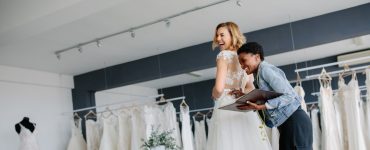 A smiling blonde woman tries on a wedding dress in a salon while another woman with a notebook and tape measure assists her.