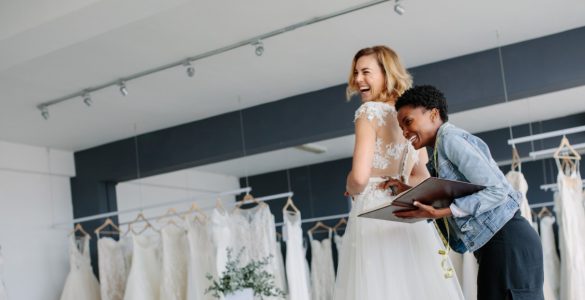 A smiling blonde woman tries on a wedding dress in a salon while another woman with a notebook and tape measure assists her.
