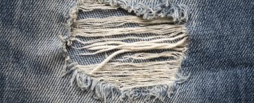 A close-up shot of light-wash denim jeans, featuring a ripped ankle with frayed threads hanging loose.