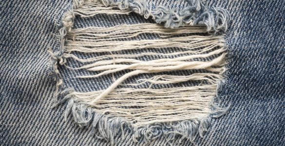 A close-up shot of light-wash denim jeans, featuring a ripped ankle with frayed threads hanging loose.