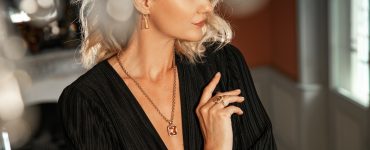 A blonde woman poses wearing a deep V-neck top with bold earrings, a pendant necklace, and a cocktail ring.