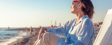 An older woman in sunglasses relaxes in a beach chair by the shore, gazing at the ocean in warm sunlight on a calm seaside day.