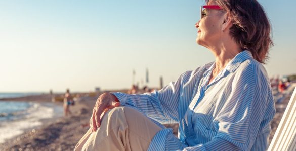 An older woman in sunglasses relaxes in a beach chair by the shore, gazing at the ocean in warm sunlight on a calm seaside day.