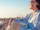 An older woman in sunglasses relaxes in a beach chair by the shore, gazing at the ocean in warm sunlight on a calm seaside day.