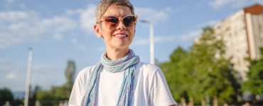 A smiling woman wearing sunglasses, a white shirt, and a colorful scarf stands outdoors on a sunny city street.