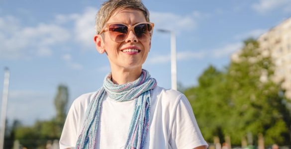 A smiling woman wearing sunglasses, a white shirt, and a colorful scarf stands outdoors on a sunny city street.