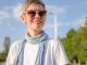 A smiling woman wearing sunglasses, a white shirt, and a colorful scarf stands outdoors on a sunny city street.