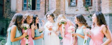 A bride in a white dress standing outdoors with several women in matching pink and blue dresses holding bouquets beside her.