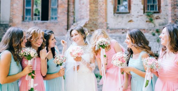 A bride in a white dress standing outdoors with several women in matching pink and blue dresses holding bouquets beside her.