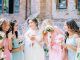 A bride in a white dress standing outdoors with several women in matching pink and blue dresses holding bouquets beside her.