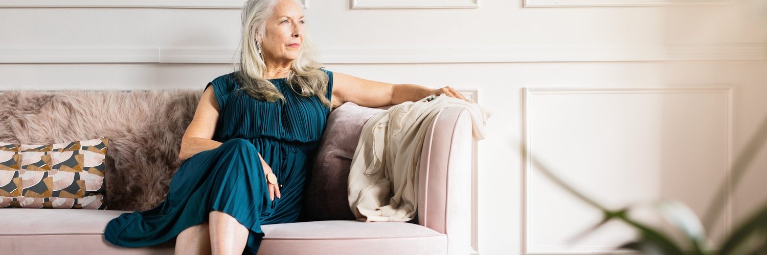 An elegant older woman in a dress sitting thoughtfully on a couch in a bright, luxurious living room.