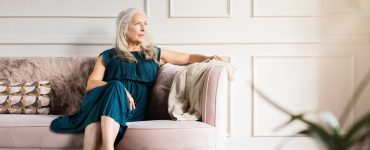 An elegant older woman in a dress sitting thoughtfully on a couch in a bright, luxurious living room.