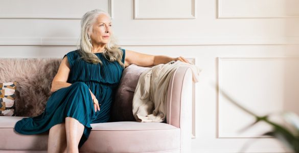 An elegant older woman in a dress sitting thoughtfully on a couch in a bright, luxurious living room.