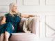 An elegant older woman in a dress sitting thoughtfully on a couch in a bright, luxurious living room.