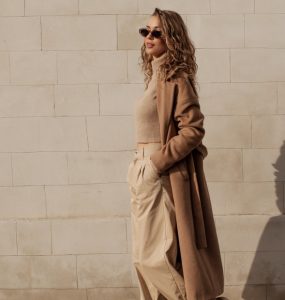 A woman wearing neutral-toned clothing and sunglasses walks beside a stone wall with her shadow cast behind her.