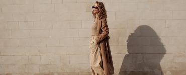A woman wearing neutral-toned clothing and sunglasses walks beside a stone wall with her shadow cast behind her.
