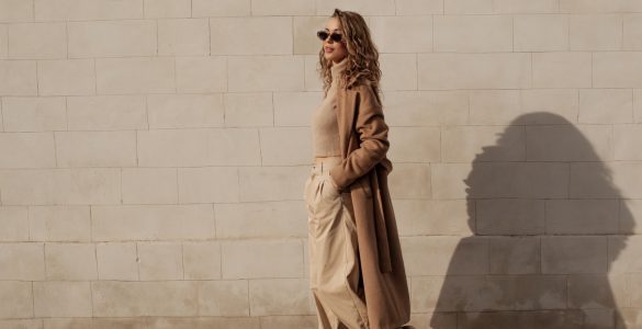 A woman wearing neutral-toned clothing and sunglasses walks beside a stone wall with her shadow cast behind her.