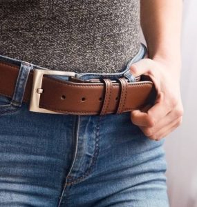 A person wearing blue jeans and a gray shirt adjusting their brown leather belt with a metal buckle near a window.