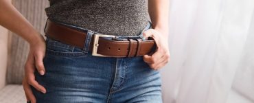 A person wearing blue jeans and a gray shirt adjusting their brown leather belt with a metal buckle near a window.
