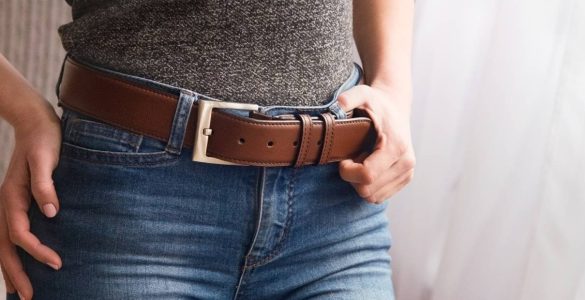 A person wearing blue jeans and a gray shirt adjusting their brown leather belt with a metal buckle near a window.