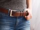 A person wearing blue jeans and a gray shirt adjusting their brown leather belt with a metal buckle near a window.