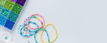 A divided craft box holding multicolored beads, bracelets, charms, scissors, and cord on a white surface.