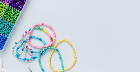 A divided craft box holding multicolored beads, bracelets, charms, scissors, and cord on a white surface.
