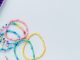 A divided craft box holding multicolored beads, bracelets, charms, scissors, and cord on a white surface.