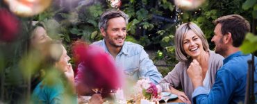 Friends laughing at a candlelit outdoor dinner table with string lights, fresh flowers, and shared plates in a garden.