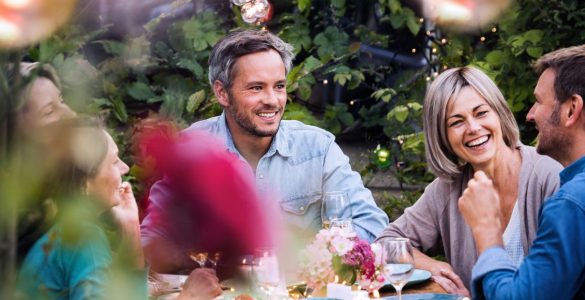 Friends laughing at a candlelit outdoor dinner table with string lights, fresh flowers, and shared plates in a garden.