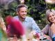 Friends laughing at a candlelit outdoor dinner table with string lights, fresh flowers, and shared plates in a garden.