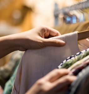 A person browsing lightweight fabric tops on wooden hangers along a clothing store rack.