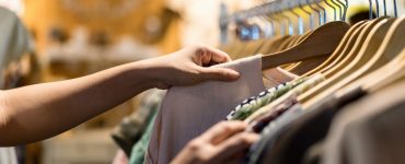 A person browsing lightweight fabric tops on wooden hangers along a clothing store rack.