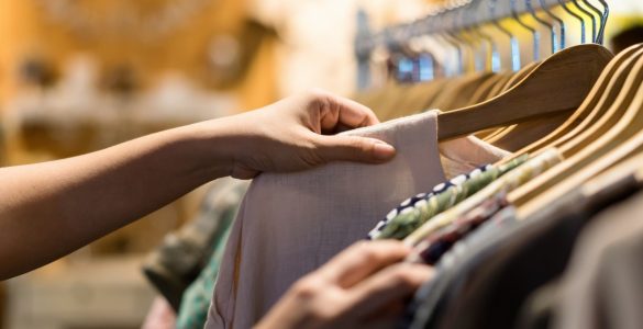 A person browsing lightweight fabric tops on wooden hangers along a clothing store rack.
