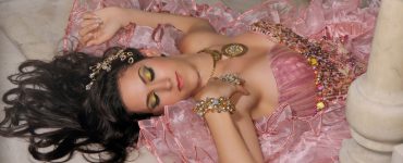 Woman in an elaborate pink ruffled gown reclines on a marble floor, wearing ornate gold jewelry and a jeweled headpiece.